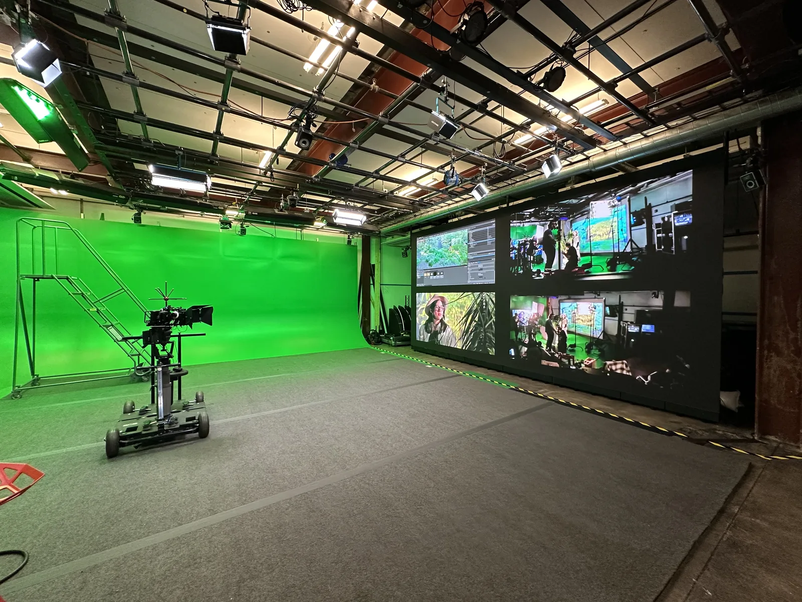 The ACE-Lab floor seen end to end: the green screen cyclorama sweeping across the left wall, a camera dolly parked at centre, and the monitor wall and control position along the right