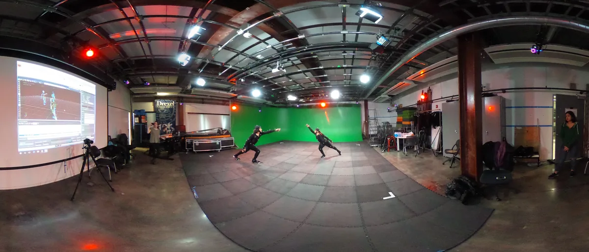 Two performers in motion capture suits mid action in the ACE-Lab volume, surrounded by tracking cameras, with a live capture preview projected on the wall behind them